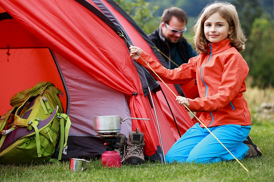 oung girl setting up a red camping tent with a backpack, cooking gear, and boots in the foreground, while an adult assists in the background at a family-friendly campground.